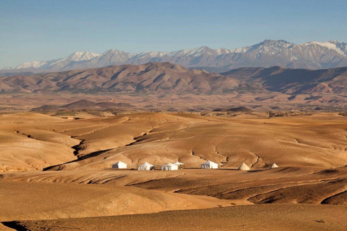 Golden hills of the Agafay desert with a tented camp and the snowy High Atlas behind